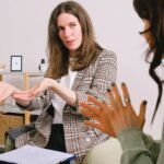 Concentrated female in casual clothes sitting with clipboard and communicating with unrecognizable colleague in light room
